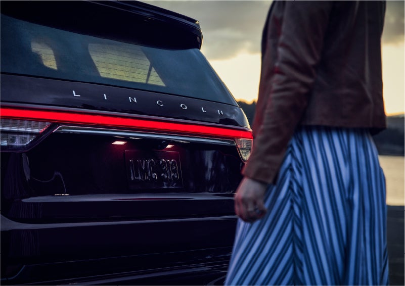 A person is shown near the rear of a 2023 Lincoln Aviator® SUV as the Lincoln Embrace illuminates the rear lights | Empire Lincoln in Abingdon VA